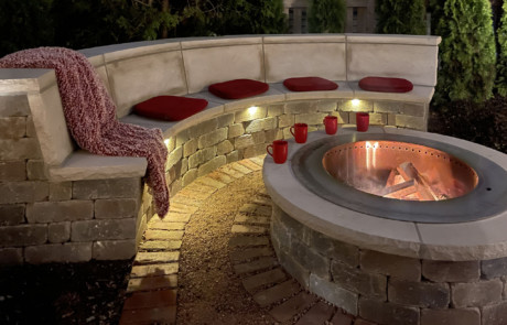 Heated modular concrete bench and smokeless wood-burning fire pit with limestone copings and under-coping lights, in rotten granite patio edged with reclaimed clay pavers